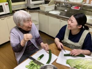 笑顔で利用者の方と台所でキュウリを切るMさんの画像